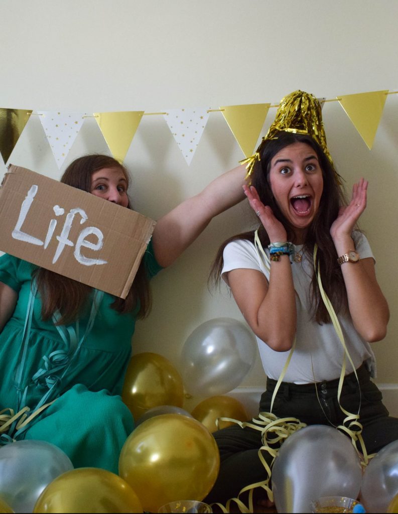 Dos chicas en una habitación decorada con artículos de fiesta, una de ellas sujeta un cartel con la palabra Life y otra está sorprendida.